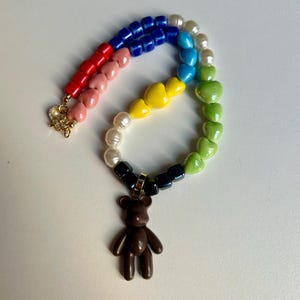 Puede incluir: Un collar de cuentas de colores con un patrón de arcoíris de cuentas en forma de corazón rojo, rosa, azul, turquesa, amarillo y verde. El collar tiene un dije de oso de peluche marrón y un cierre dorado.