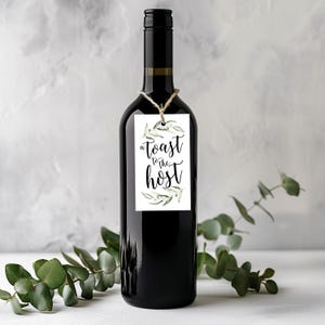 May include: A dark glass wine bottle with a rectangular white paper tag tied around the neck with twine. The tag features green watercolour olive branch illustrations and the black printed text A toast to the host in a decorative script font.