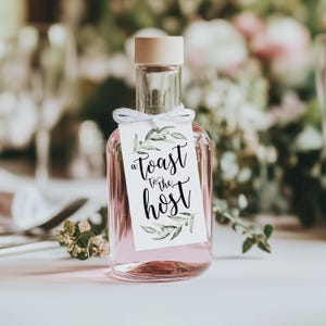 May include: A clear glass bottle filled with a light pink liquid, topped with a light wood stopper. A white rectangular tag is tied to the neck with white string, featuring green watercolour leaf illustrations and the black text A TOAST TO THE HOST.