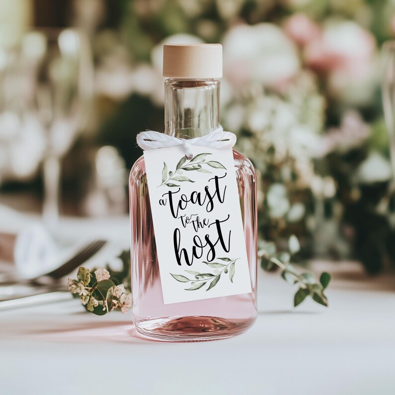 May include: A clear glass bottle filled with a light pink liquid, topped with a light wood stopper. A white rectangular tag is tied to the neck with white string, featuring green watercolour leaf illustrations and the black text A TOAST TO THE HOST.