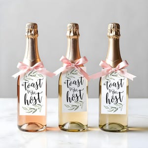 May include: Three glass bottles with gold foil necks, each featuring a light pink satin ribbon tied around the neck. A rectangular white tag hangs from each ribbon, printed with green olive branch illustrations and the text A toast to the host.