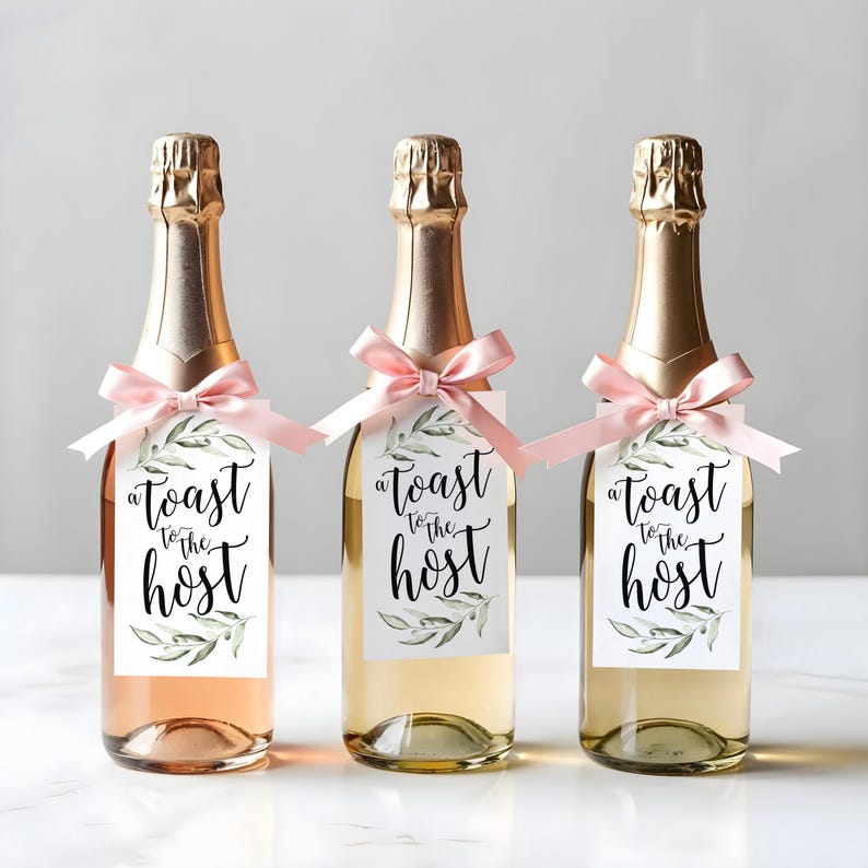May include: Three glass bottles with gold foil necks, each featuring a light pink satin ribbon tied around the neck. A rectangular white tag hangs from each ribbon, printed with green olive branch illustrations and the text A toast to the host.