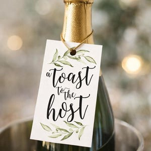 May include: A rectangular white gift tag hanging from a gold string on a champagne bottle neck. The tag features green watercolour olive branch illustrations at the top and bottom with the black cursive text A toast to the host in the centre.