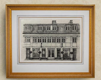 Puede incluir: Dibujo a pluma enmarcado de un edificio con múltiples ventanas y tejado de tejas. La obra de arte está en un marco dorado con un passe-partout blanco. El edificio tiene una fachada detallada con elementos arquitectónicos.