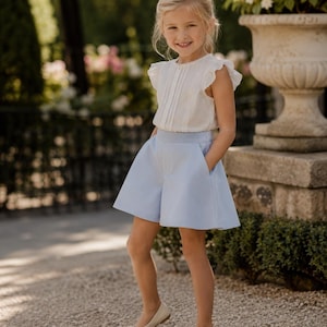 Könnte beinhalten: Ein junges Mädchen trägt ein weißes Oberteil mit Rüschen und hellblaue Shorts. Dazu trägt sie cremefarbene Ballerinas. Das Outfit ist für einen warmen Tag geeignet. Der Hintergrund umfasst Grün und einen Steintopf.