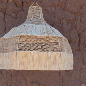 May include: A woven pendant light fixture with a natural, beige color. The lamp has a conical shape with a fringed bottom edge and is suspended from a woven cord. The background is a textured, reddish-brown wall.