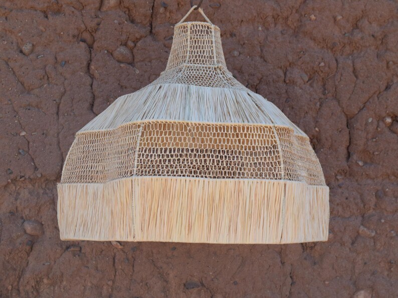 May include: A natural woven pendant light fixture. The light shade is made of light beige woven material with a fringed bottom. The light fixture is hanging against a textured brown wall.