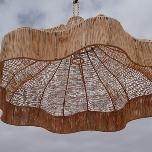 May include: A large, woven pendant light fixture with a natural, light brown color. The shade has a wavy, scalloped edge and a woven interior. The light hangs from a woven cord against a cloudy sky.