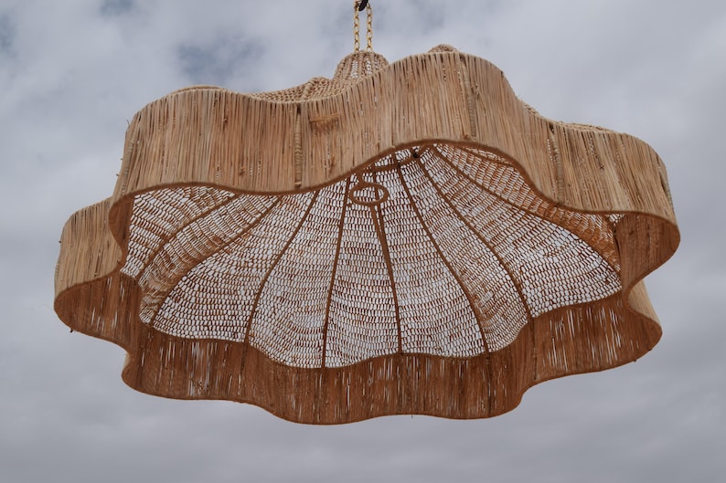 May include: A large, woven pendant light fixture with a natural, light brown color. The shade has a wavy, scalloped edge and a woven interior. The light hangs from a woven cord against a cloudy sky.