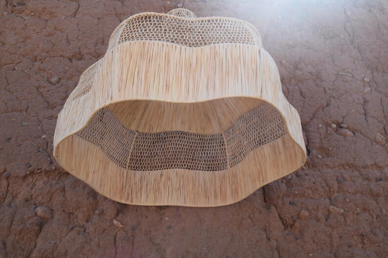 May include: A natural-toned, woven lampshade with a unique, layered design. The lampshade features a combination of open weave and fringed textures, creating a decorative lighting element. The neutral color and organic materials suggest a bohemian or rustic aesthetic.