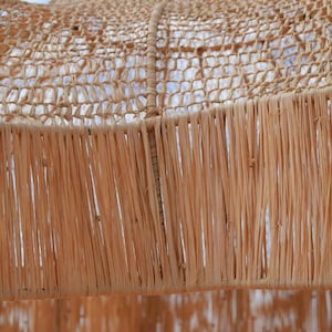 May include: Close-up of a woven lampshade with a natural, light brown color. The lampshade has a netted top and a fringed bottom, creating a textured and airy appearance. The design suggests a bohemian or coastal decor style.