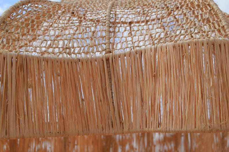 May include: Close-up of a woven lampshade with a natural, light brown color. The lampshade has a netted top and a fringed bottom, creating a textured and airy appearance. The design suggests a bohemian or coastal decor style.