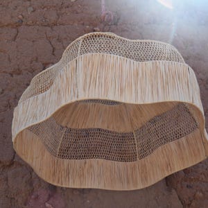 May include: A woven, natural fiber lampshade with a unique, wavy design. The lampshade features a fringed lower edge and a woven, openwork pattern. The neutral color and organic texture create a warm, inviting aesthetic. The lampshade is designed to be hung from the ceiling.