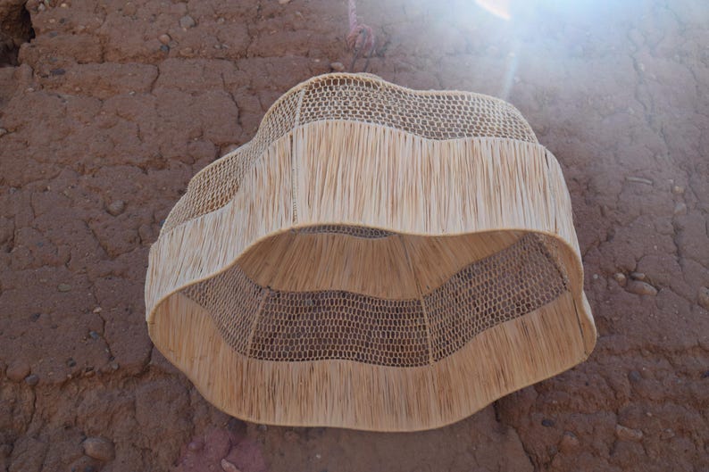 May include: A woven, natural fiber lampshade with a unique, wavy design. The lampshade features a fringed lower edge and a woven, openwork pattern. The neutral color and organic texture create a warm, inviting aesthetic. The lampshade is designed to be hung from the ceiling.