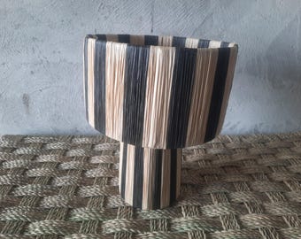 Puede incluir: Un macetero cilíndrico tejido en negro y natural con una parte superior ancha y una base estrecha. El macetero está hecho de tiras tejidas de material natural y material negro.