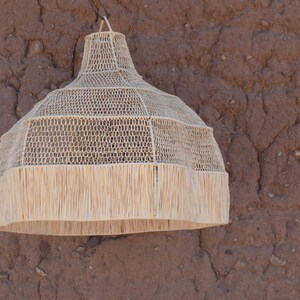 May include: A beige woven pendant light fixture with a fringed bottom. The light has a bell shape and is suspended against a textured, reddish-brown wall. The light fixture is made of natural materials.