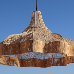 May include: A large, woven pendant light fixture with a natural, beige color. The light has a bell-shaped design with a wide, scalloped bottom and a textured, open weave. The fixture hangs from a dark cord against a clear blue sky.