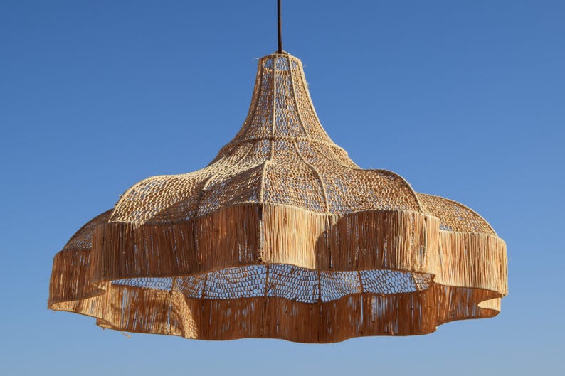 May include: A large, woven pendant light fixture with a natural, beige color. The light has a bell-shaped design with a wide, scalloped bottom and a textured, open weave. The fixture hangs from a dark cord against a clear blue sky.