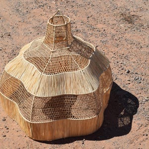 May include: A handcrafted, beige woven basket with a unique, bulbous shape. The basket features a flared top and a textured, layered design. The natural fibers create a rustic aesthetic, perfect for home decor or storage.