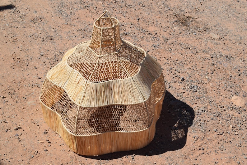 May include: A handcrafted, beige woven basket with a unique, bulbous shape. The basket features a flared top and a textured, layered design. The natural fibers create a rustic aesthetic, perfect for home decor or storage.