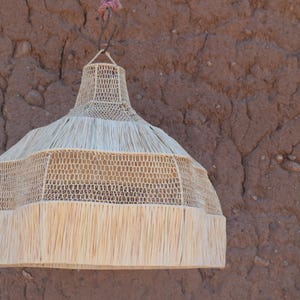 May include: A woven pendant light fixture with a natural, beige color. The light has a bell shape with a fringed bottom and is suspended from a brown wall. The light fixture is made of woven material and has a decorative design.