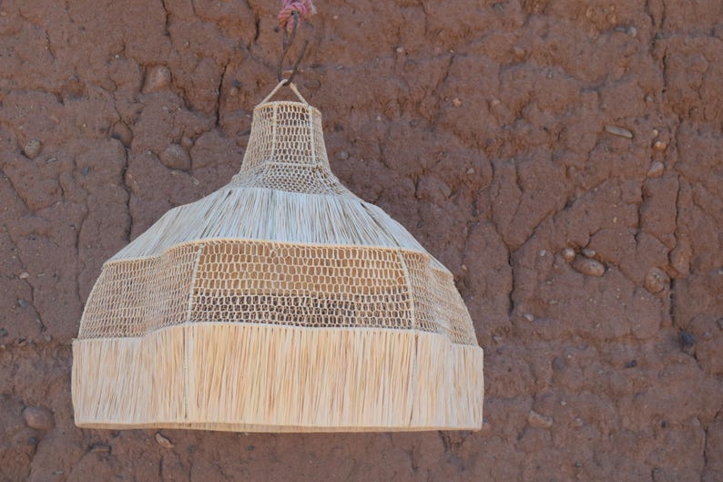 May include: A woven pendant light fixture with a natural, beige color. The light has a bell shape with a fringed bottom and is suspended from a brown wall. The light fixture is made of woven material and has a decorative design.
