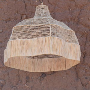 May include: A woven pendant light fixture with a natural, beige color. The lamp has a bell-shaped design with a fringed bottom. The light fixture is hanging against a textured, brown wall.