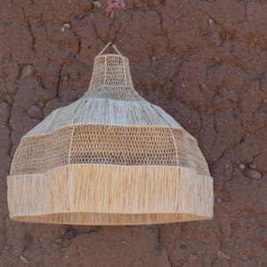 May include: A woven pendant light fixture with a natural, beige color. The lamp has a conical shape with a fringed bottom edge. The fixture is suspended from a brown wall.