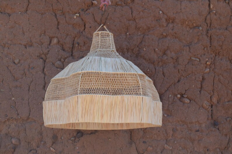 May include: A woven pendant light fixture with a natural, beige color. The lamp has a conical shape with a fringed bottom edge. The fixture is suspended from a brown wall.