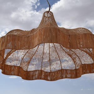 May include: A woven pendant light fixture with a natural, light brown color. The shade has a scalloped edge and a netted interior, creating a decorative and airy design. The light hangs from a black cord against a cloudy sky.