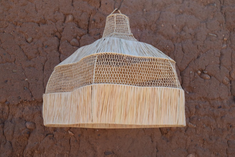 May include: A natural woven pendant light fixture with a conical top and a flared, fringed bottom. The light is made of light tan woven material and hangs against a textured brown wall.