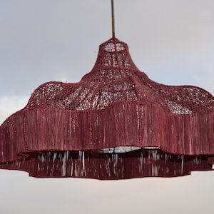 Puede incluir: Una lámpara colgante tejida de color burdeos. La pantalla tiene un diseño en capas con un borde festoneado y una textura de tejido abierto. La luz cuelga de un solo cable contra un cielo nublado.