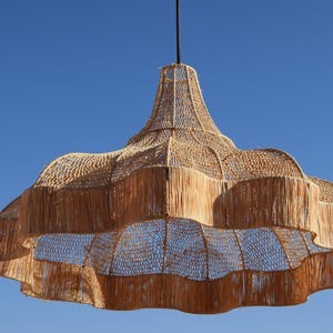May include: A large, woven pendant light fixture with a natural, light brown color. The fixture has a layered, scalloped design and hangs from a black cord against a clear blue sky. The light is made of natural materials.