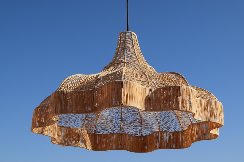 May include: A large, woven pendant light fixture with a natural, light brown color. The fixture has a layered, scalloped design and hangs from a black cord against a clear blue sky. The light is made of natural materials.