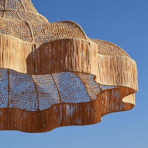 May include: A woven, tiered pendant light fixture with a natural, beige color. The light has a fringed edge and a woven, open-weave design. The background is a clear, bright blue sky.
