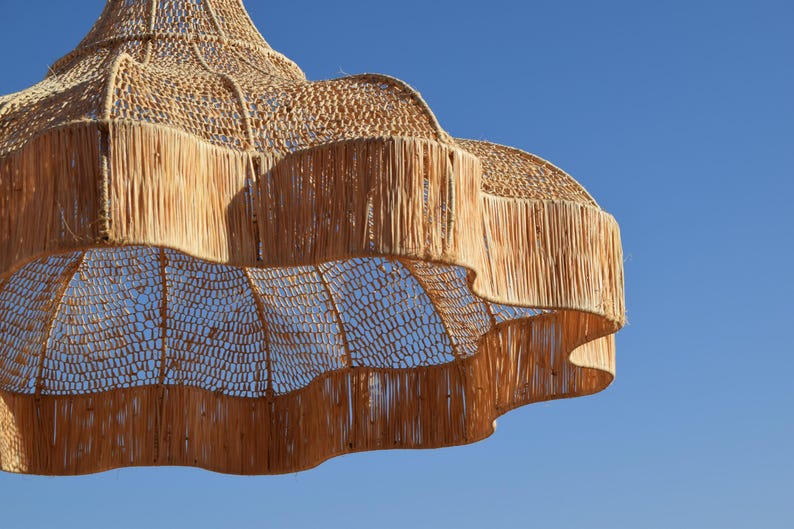 May include: A woven, tiered pendant light fixture with a natural, beige color. The light has a fringed edge and a woven, open-weave design. The background is a clear, bright blue sky.