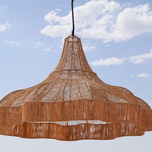 May include: A woven pendant light fixture with a natural brown color. The light has a wide, tiered shade with a fringed edge and a bell-shaped top. It hangs from a black cord against a blue sky with clouds.