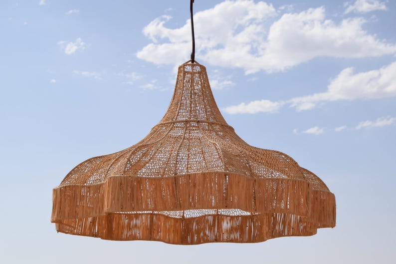 May include: A woven pendant light fixture with a natural brown color. The light has a wide, tiered shade with a fringed edge and a bell-shaped top. It hangs from a black cord against a blue sky with clouds.