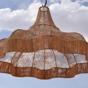 May include: A large, woven pendant light fixture with a natural, light brown color. The fixture has a scalloped edge and a bell-shaped top, suspended against a blue sky with fluffy white clouds. The light is made of woven material.