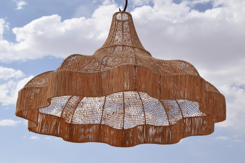 May include: A large, woven pendant light fixture with a natural, light brown color. The fixture has a scalloped edge and a bell-shaped top, suspended against a blue sky with fluffy white clouds. The light is made of woven material.