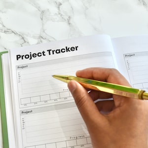 May include: A close-up of an open notebook with the title "Project Tracker" written in black on the top of the page. The notebook has a green spine and is lying on a white marble surface. A hand is holding a gold pen and writing in the notebook.