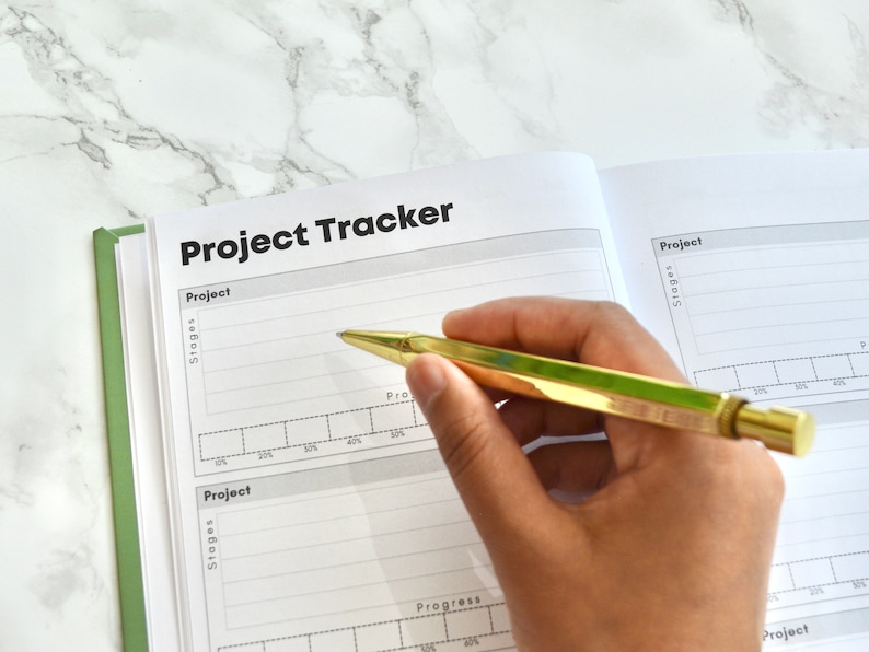 May include: A close-up of an open notebook with the title "Project Tracker" written in black on the top of the page. The notebook has a green spine and is lying on a white marble surface. A hand is holding a gold pen and writing in the notebook.