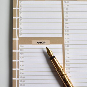May include: A gold-toned fountain pen with a decorative design on a brown and white planner with a section labeled "MORNING". The planner has a grid of boxes and times listed in increments of 30 minutes.