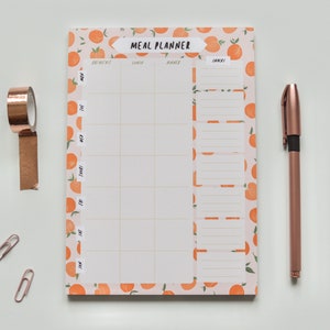 May include: A peach-patterned meal planner with a white background and a grid for writing in meals for each day of the week. The planner has sections for breakfast, lunch, dinner, and snacks. The planner is on a white surface with a roll of copper tape and two paper clips.