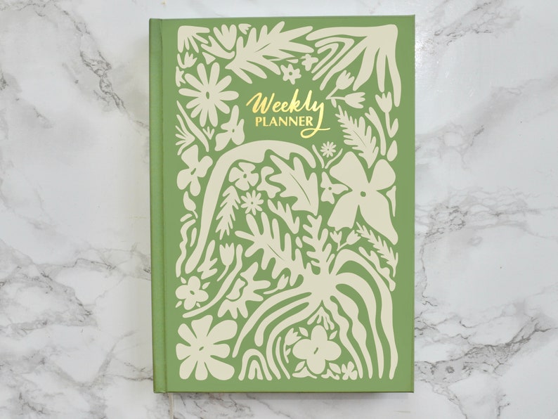 May include: A green weekly planner with a white floral design and the text "Weekly Planner" in gold lettering.