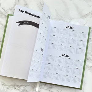 May include: An open green planner with the words "My Roadmap" on the cover. The planner has a monthly calendar for 2025 and 2026.