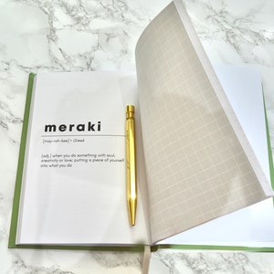 May include: An open journal with a green cover and white pages. The word "meraki" is printed on the page, along with its definition and pronunciation. A gold pen is lying on the open page.