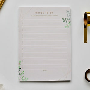 Puede incluir: Un bloc de notas blanco con un diseño floral y el texto "THINGS TO DO" y "IT ALWAYS SEEMS IMPOSSIBLE UNTIL IT IS DONE". El bloc de notas tiene una sección rayada para escribir y una sección para notas.