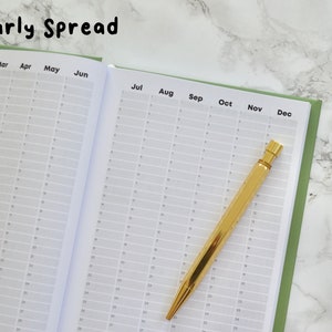 May include: A green and white yearly planner with a gold pen lying on top of the open pages. The planner has a grid layout with months labeled from January to December.