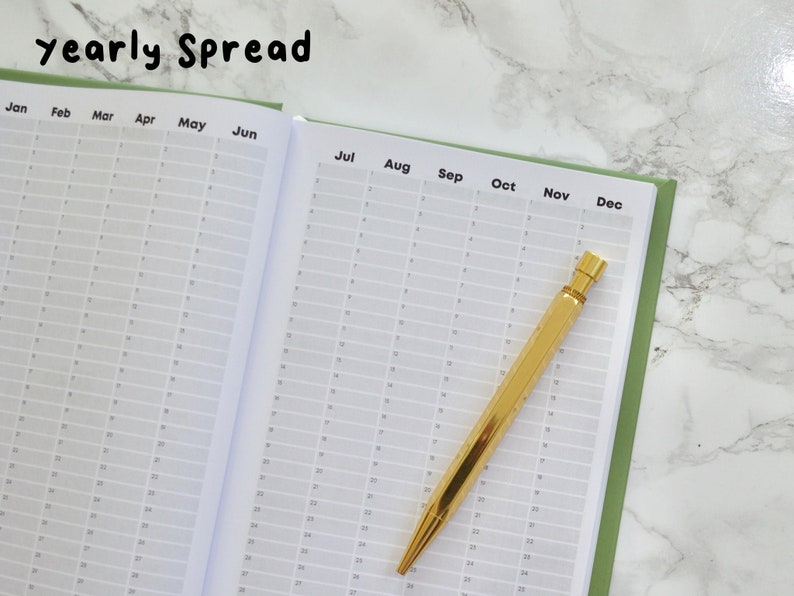 May include: A green and white yearly planner with a gold pen lying on top of the open pages. The planner has a grid layout with months labeled from January to December.